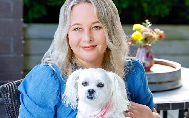 Blonde woman in blue blouse sitting outdoors, holding a small white dog with a pink scarf.