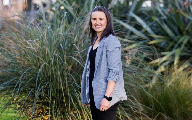 Bernadine Bezuidenhout in a grey blazer standing in front of some green plants
