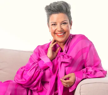 Paula Bennett dressed in a hot pink outfit leans against the arm of a blush pink sofa her other hand grazes her neck