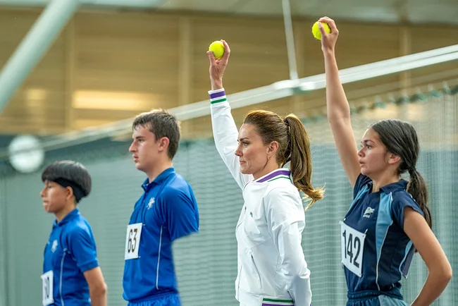 Four people, including a woman in white, hold tennis balls up in a sports facility, wearing athletic attire.