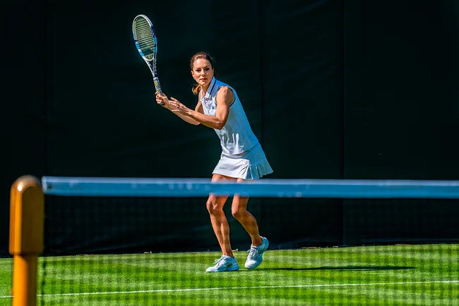 A woman in a white tennis outfit holds a racket, preparing to hit a ball on a grass court.
