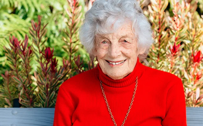 Elderly woman with white hair in a red sweater smiles, sitting on a bench with greenery in the background.