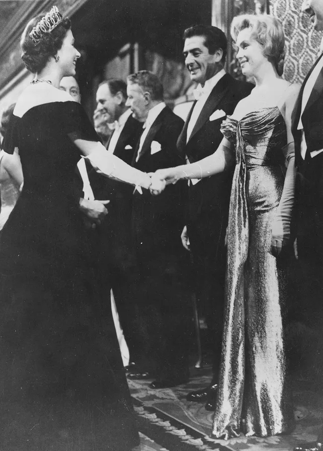 A woman in a tiara shakes hands with a woman in a sparkling gown at an elegant event, surrounded by men in tuxedos.