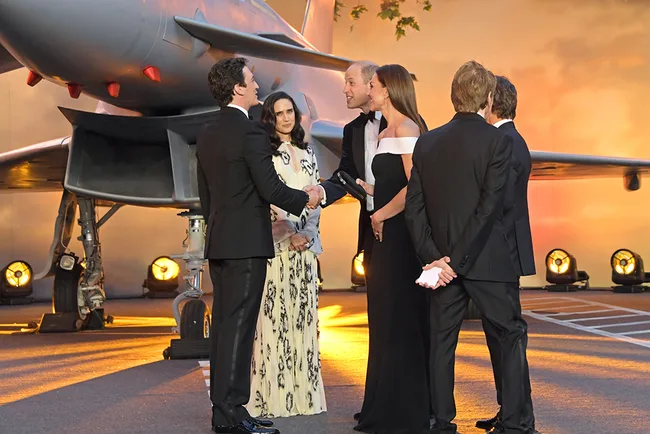 Group of people, including figures in suits and dresses, stand near a jet on a brightly lit evening.