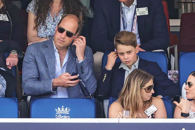 A man and a boy in suits sit in a stadium, adjusting earpieces. Two women sit in front, talking and drinking.