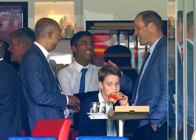 A young boy eating pizza, surrounded by three adults in suits, smiling and talking in a room.