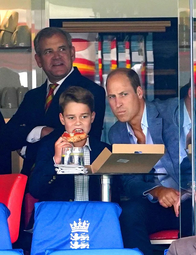 Boy eating pizza with two men at a sports event, seated at a table with a box, blue chair covers with a crown logo.