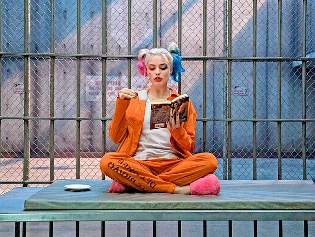 Harley Quinn in an orange jumpsuit, sitting on a prison bed with a book and a cup, in "Suicide Squad" movie scene.
