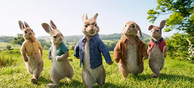 Five animated rabbits wearing clothes walk on a grassy hill, with trees and blue sky in the background.