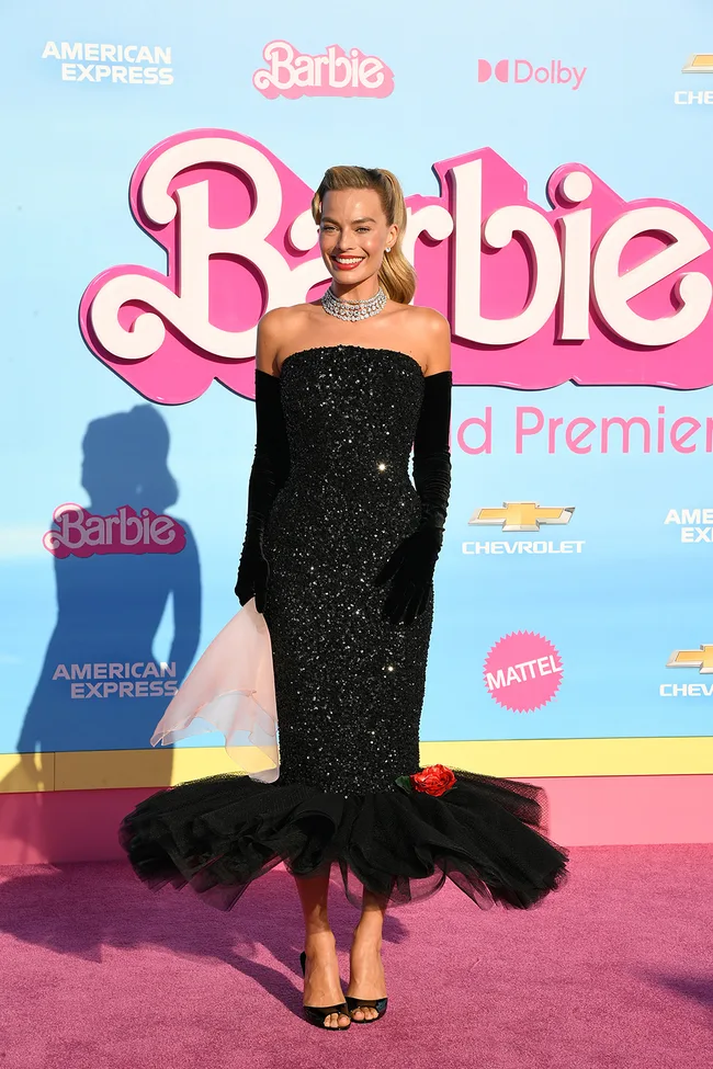 Actress in a sparkling black dress poses at the Barbie movie premiere on a pink carpet with a blue background.