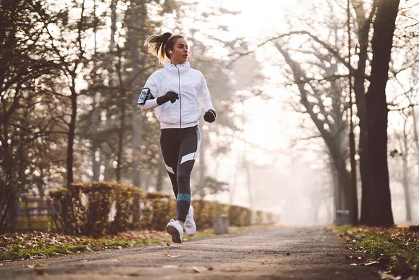 Woman jogging on a tree-lined path in a park, wearing sportswear and listening to music.