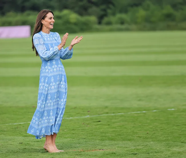 A woman in a blue patterned dress clapping on a grass field, smiling with trees in the background.