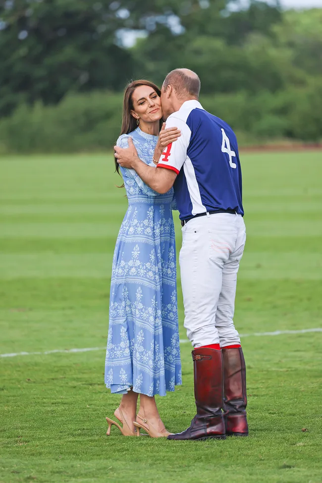 A woman in a blue dress hugs a man in a polo uniform with tall boots on a green field.