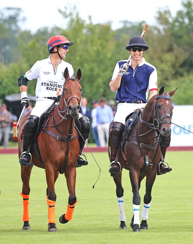 Two polo players on horseback, one in a white jersey and red helmet, the other in blue, smiling while riding on a green field.