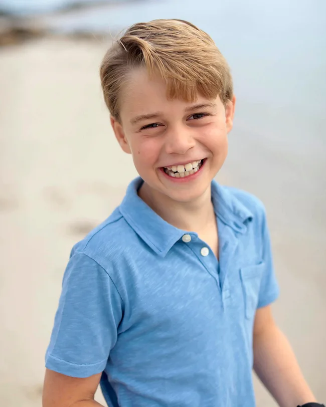 A child with light hair smiling, wearing a blue shirt, stands on a beach with a blurred background.