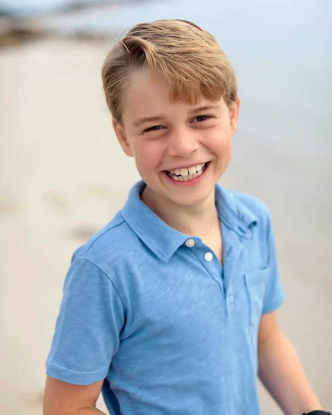 A child with light hair smiling, wearing a blue shirt, stands on a beach with a blurred background.