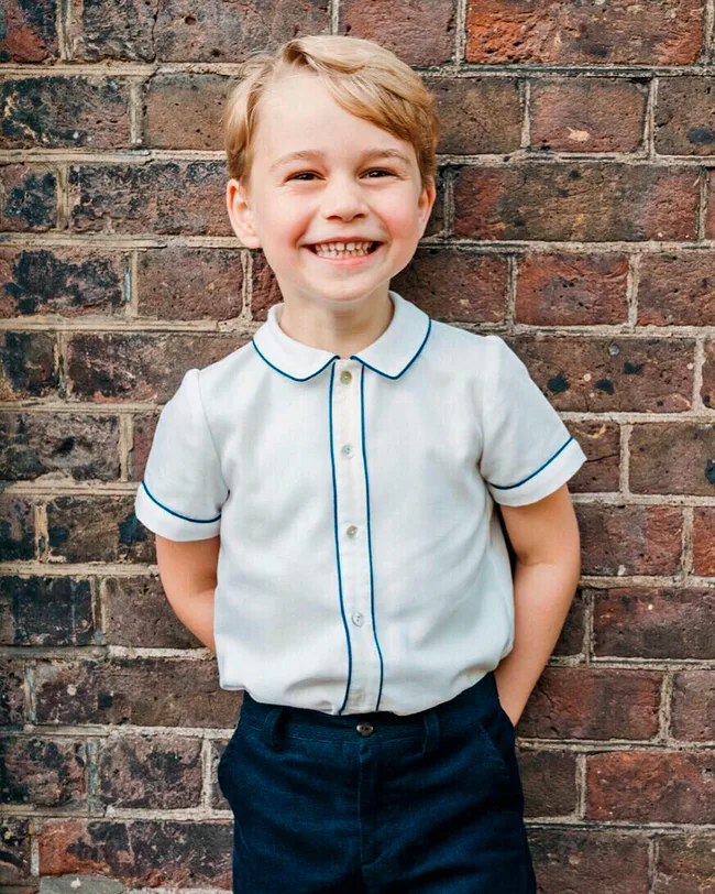 Young boy smiling brightly in a white shirt and blue shorts, standing against a brick wall.