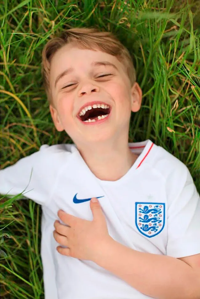 A young boy wearing an England soccer jersey laughs joyfully while lying on grass.