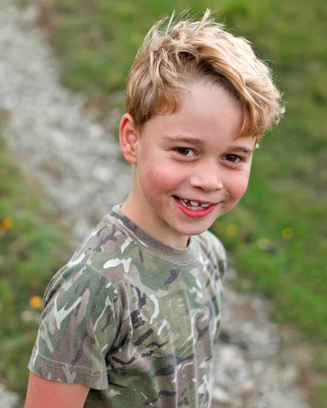 A young boy with blonde hair wearing a camouflage t-shirt smiles outdoors.