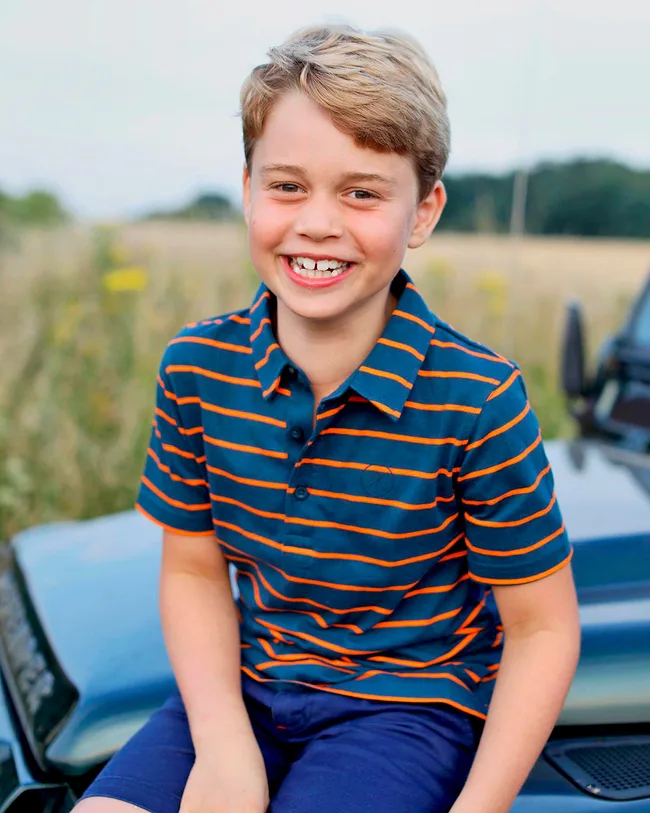 A child with blonde hair smiles, sitting outdoors on a vehicle, wearing a blue and orange striped shirt.