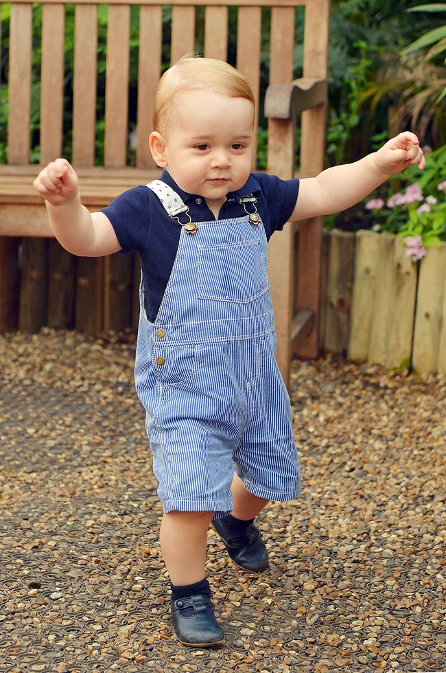 Toddler in blue overalls taking steps on a gravel path near a wooden bench and garden flowers.