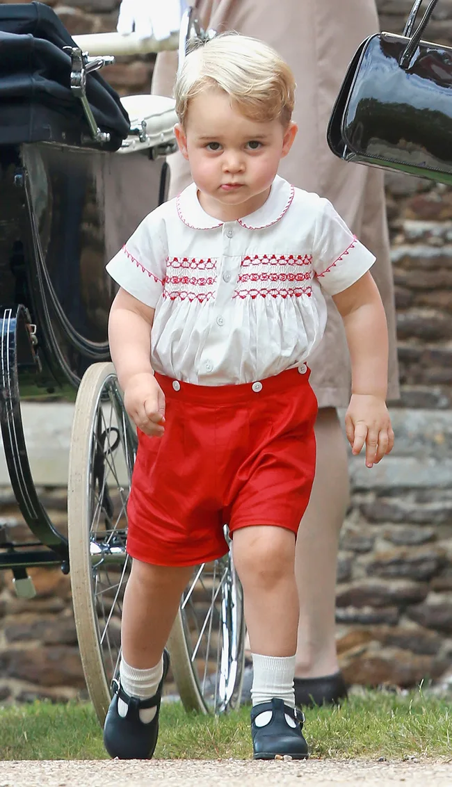 Toddler in white shirt with red embroidery and shorts, walking near a black pram outdoors.