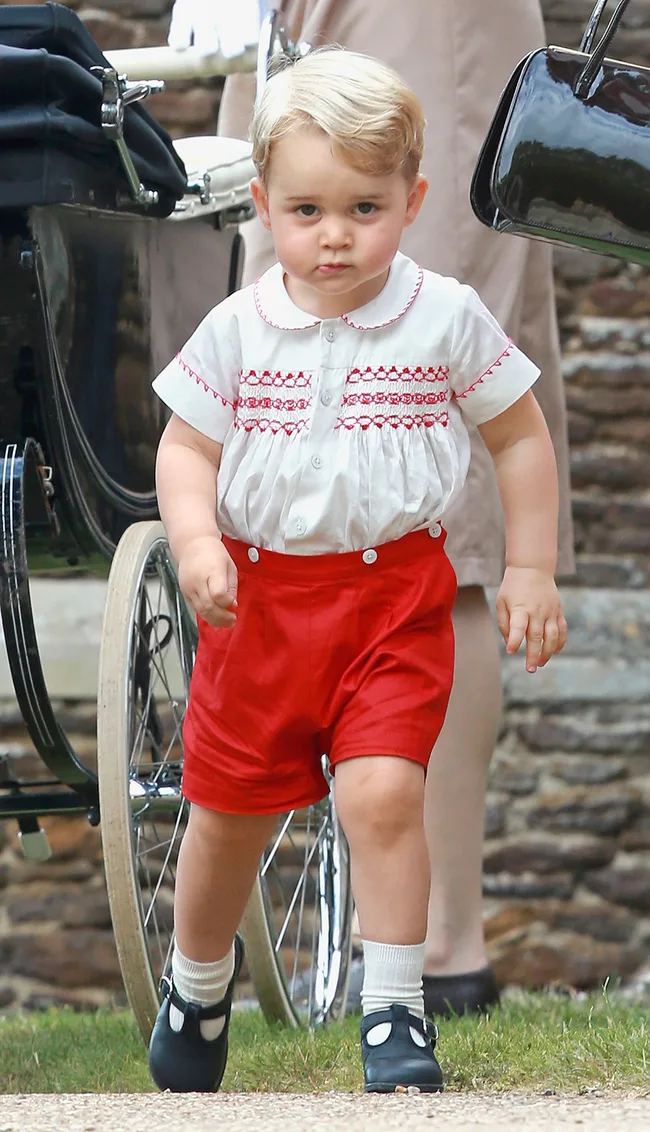 Toddler in white shirt with red embroidery and shorts, walking near a black pram outdoors.