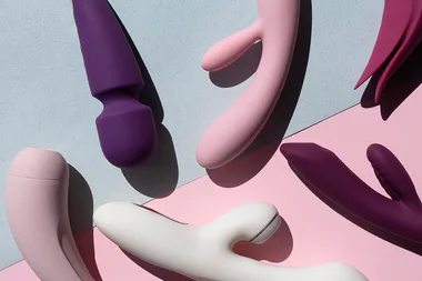 Various vibrators in shades of purple, pink, and white are arranged on a light blue and pink background.