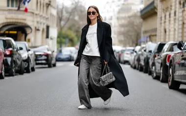 A person walks on a city street wearing sunglasses, a long black coat, white t-shirt, gray jeans, and holding a handbag.