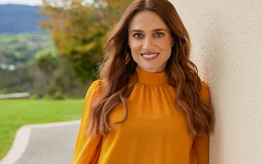 A woman with long brown hair in an orange top smiles while standing outside against a light-colored wall.