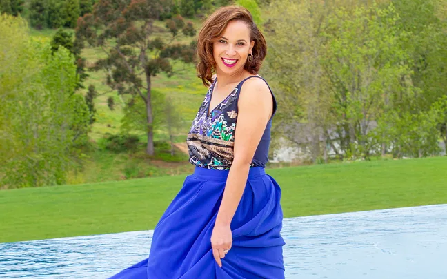 A woman in a colorful top and blue skirt smiles by a poolside with a green park in the background.