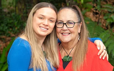 Two women smiling in a close embrace, one wearing a blue top and the other a red top, with foliage in the background.