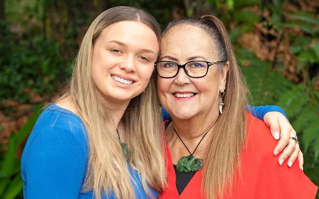 Two women smiling in a close embrace, one wearing a blue top and the other a red top, with foliage in the background.