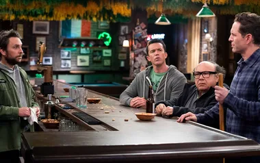 Four men standing at a cluttered bar in a dimly lit pub with Irish decorations.