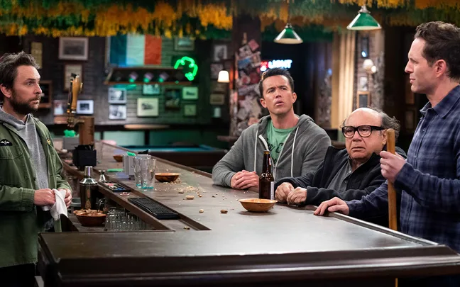 Four men standing at a cluttered bar in a dimly lit pub with Irish decorations.