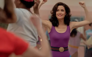 Woman in a purple leotard doing an aerobic exercise class, smiling with arms flexed.