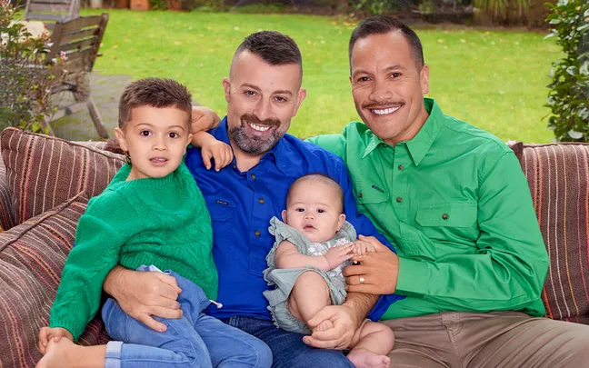 Two smiling adults with a toddler and a baby sitting on a couch in a garden setting.