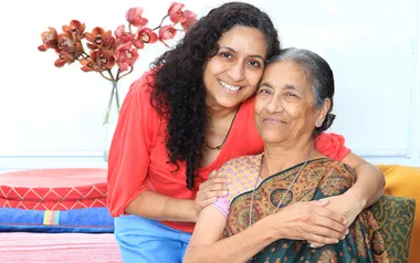 Mother and daughter smiling, daughter in red top hugging mother in colorful saree, seated indoors with flowers in background.