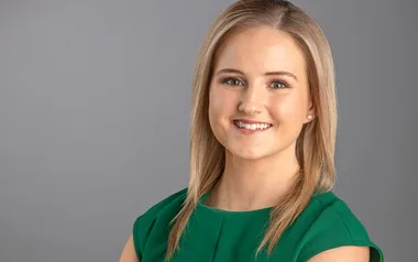 A woman with straight blonde hair smiles, wearing a green dress against a gray background.
