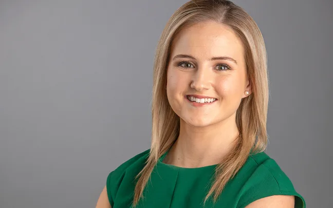 A woman with straight blonde hair smiles, wearing a green dress against a gray background.
