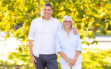 Man and woman smiling outdoors, standing in front of sunlit trees, wearing light-colored shirts.