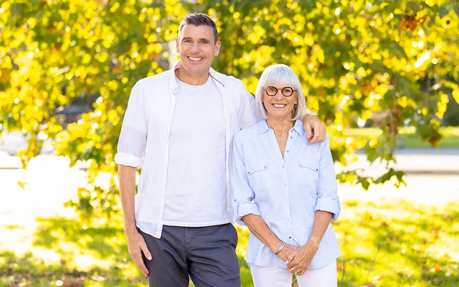 Man and woman smiling outdoors, standing in front of sunlit trees, wearing light-colored shirts.