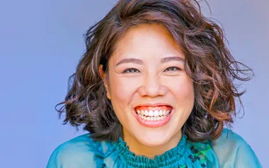 A person with wavy hair smiling broadly, wearing a blue top against a light blue background.
