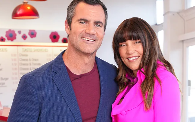 Man in blue blazer and woman in pink jacket smiling in vibrant setting with floral decor.