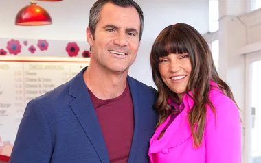 Man in blue blazer and woman in pink jacket smiling in vibrant setting with floral decor.