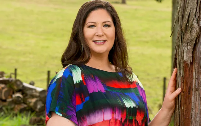 A woman in a colorful blouse stands by a tree in a grassy field, smiling at the camera.