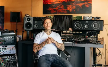 Tattooed man wearing a hat and white shirt sits in a music studio with audio equipment and speakers.