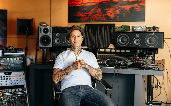 Tattooed man wearing a hat and white shirt sits in a music studio with audio equipment and speakers.