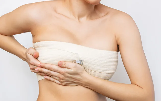 A person wearing a beige compression bandage around the chest, adjusting it with hands, against a plain background.