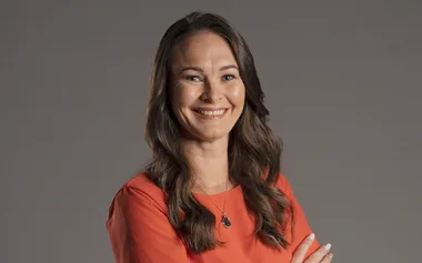 A woman with long hair wearing a red top smiles, arms crossed. Gray background.
